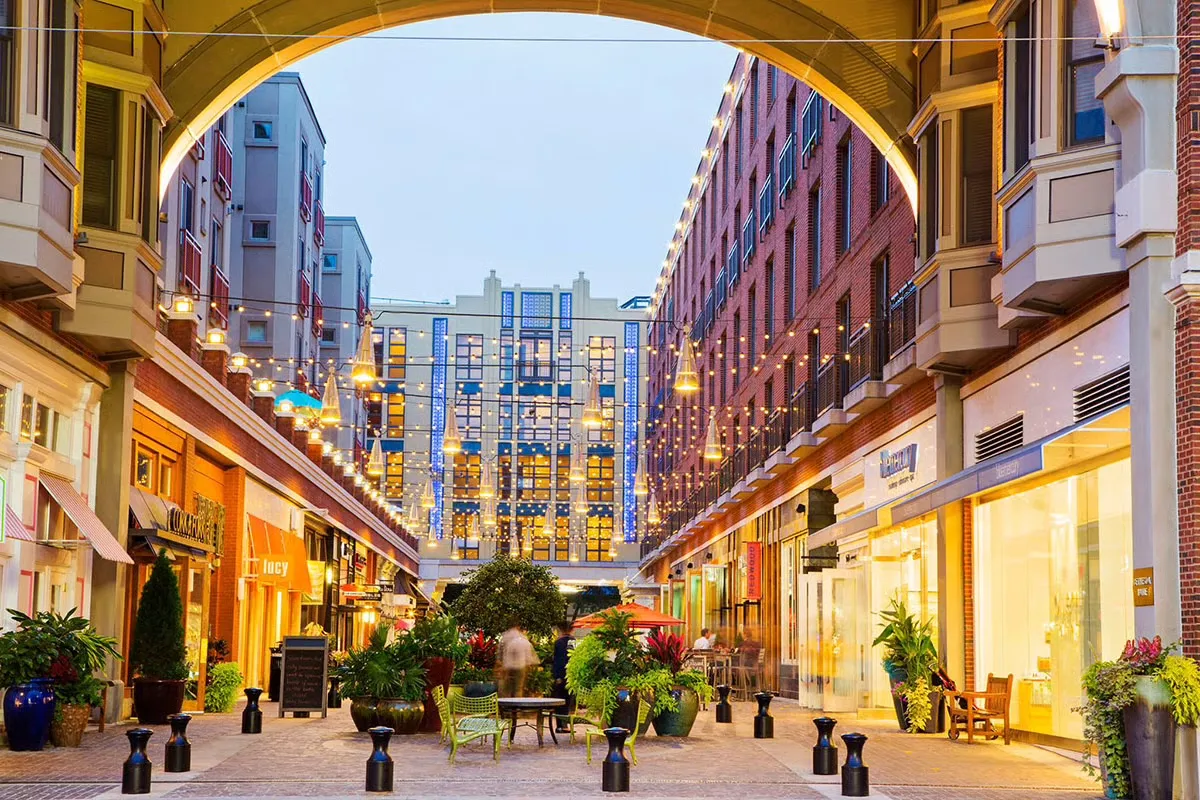 Bethesda Lane at dusk, seen from beneath its arch — string lights and cone lamps over the cobbled lane, shopfronts and planters on either side, and brick apartments above